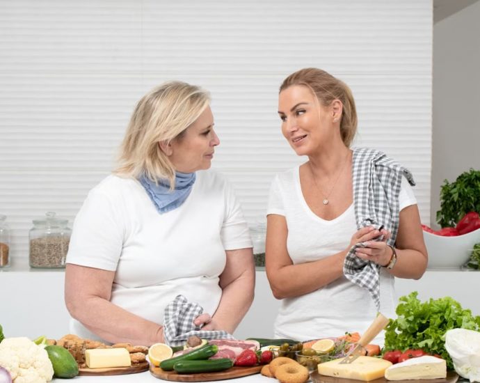 Two women in a kitchen preparing fresh vegetables and cheese with olive oil and spices.