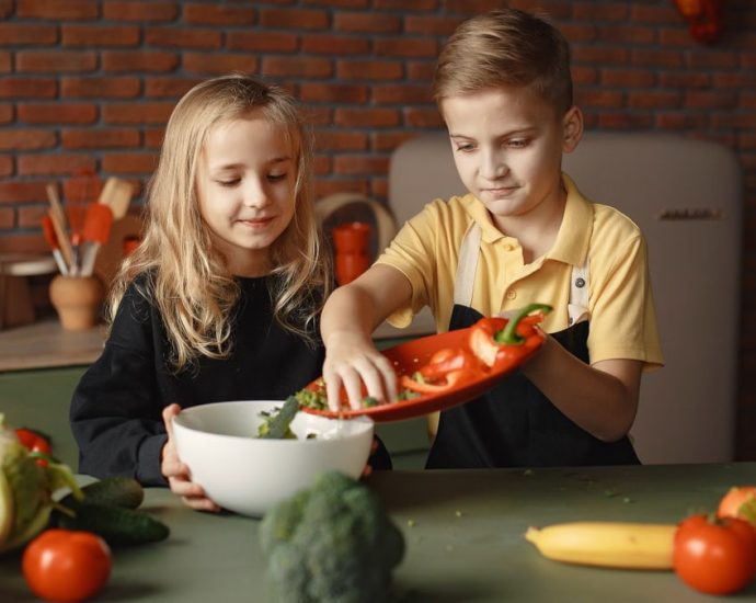 Little girl and boy mixing cutted pepper with other fresh ingredients for salad in big bowl while cooking at table with various fresh vegetables and fruits